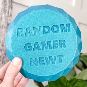 Blue geometric object with 'RANDOM GAMER NEWT' text held by a hand against a blurred natural background